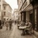 Cobblestone street in a historic European town; pedestrians in mid-20th-century attire walk by while a couple sits at a table outside a camera shop.