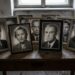 Framed black-and-white portraits of elderly people arranged on a worn wooden table in a dim room by a window.