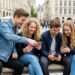Four teenagers sit on a stone bench in a cobblestone city square, smiling as they look at their phones together.