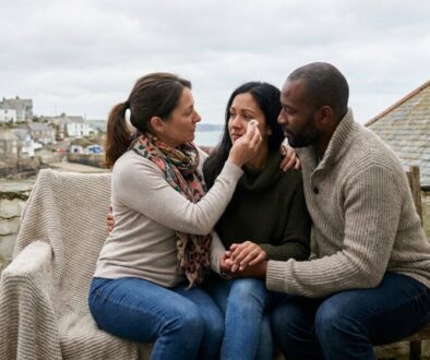 Three friends sit on a bench by a stone wall; one woman gently touches the other's face as she applies makeup, while a man watches supportively in the background.