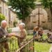 Elderly couple exchanges a loaf of bread in a cobblestone village square, with a rose garden nearby.