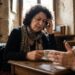 Older woman with a patterned scarf sits at a wooden table in a stone cafe, touching her temple while holding a teacup.