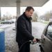 Man in a dark jacket refuels a silver car at a gas station, holding the nozzle near the fuel cap.