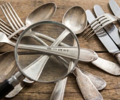 A pile of vintage silverware (forks, spoons, knives) scattered on a wooden table with a magnifying glass over them.