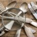 A pile of vintage silverware (forks, spoons, knives) scattered on a wooden table with a magnifying glass over them.