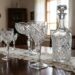 Crystal decanter and matching etched glasses arranged on a wooden dining table with a lace runners, in a sunlit room.