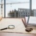 Architectural blueprints on a wooden desk with a magnifying glass, a padlock, and a gavel beside a stack of documents; construction site visible outside window.