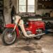 Vintage red-and-cream motorcycle parked in a dusty, cluttered workshop with tools on shelves nearby.