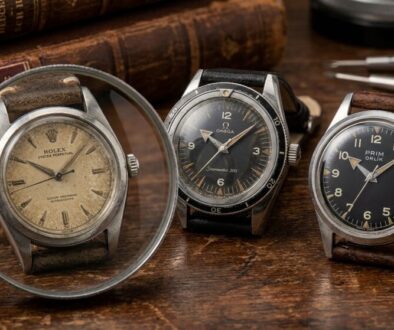 Three vintage wristwatches sit on a worn wood surface, with a magnifying glass and antique books in the background, suggestive of a collector's desk.