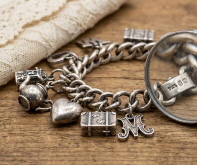 Silver charm bracelet with heart, luggage charm, and an ornate M charm on a wooden surface, with a magnifying glass nearby and beige lace in the background