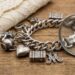 Silver charm bracelet with heart, luggage charm, and an ornate M charm on a wooden surface, with a magnifying glass nearby and beige lace in the background