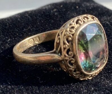 Antique gold ring with intricate scrollwork filigree around a large oval multi-color gemstone on black velvet backdrop.