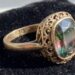 Antique gold ring with intricate scrollwork filigree around a large oval multi-color gemstone on black velvet backdrop.