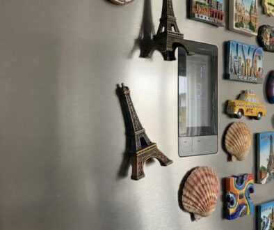 Fridge door covered in travel magnets and seashells around a vertical digital display.