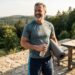 Smiling man in a athletic shirt holds a water bottle and rolled yoga mat on a stone patio with a forested hillside backdrop.