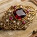 Intricately designed gold brooch with a large red gemstone surrounded by pearls, placed in a vintage box liner.