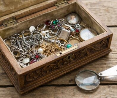Open vintage jewelry box on a wooden table, spilling silver chains, rings, and gemstone accessories; a magnifying glass rests beside it.
