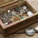 Open vintage jewelry box on a wooden table, spilling silver chains, rings, and gemstone accessories; a magnifying glass rests beside it.