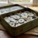 Rusty green metal tin filled with white buttons of various sizes on a wooden table, with beige fabric underneath.