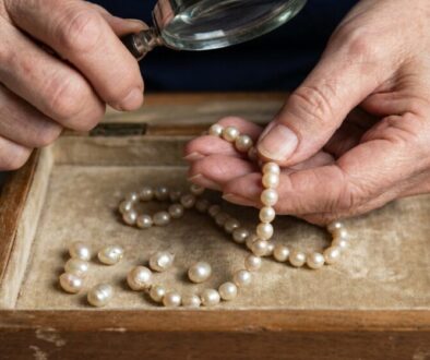 Older hands stringing or inspecting a pearl necklace with a magnifying glass over a wooden tray of pearls.