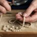 Older hands stringing or inspecting a pearl necklace with a magnifying glass over a wooden tray of pearls.
