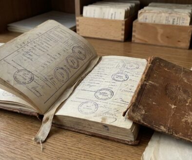 Open antique ledger with handwritten entries and circular stamps on a wooden desk, alongside a worn brown notebook and a magnifying glass.
