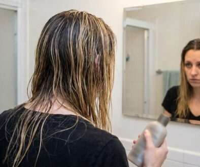Person with wet hair seen from behind in a bathroom, holding a shampoo bottle and looking at their reflection in the mirror.