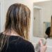Person with wet hair seen from behind in a bathroom, holding a shampoo bottle and looking at their reflection in the mirror.