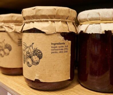 Row of jam jars on a wooden shelf with kraft labels and twine; center jar shows an 'Ingredients' list (sugar, water, fruit concentrate, pectin, citric acid).