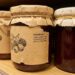 Row of jam jars on a wooden shelf with kraft labels and twine; center jar shows an 'Ingredients' list (sugar, water, fruit concentrate, pectin, citric acid).