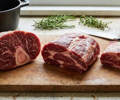 Three raw beef steaks on a wooden cutting board, with a knife and sprigs of rosemary and thyme in the background.