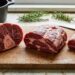 Three raw beef steaks on a wooden cutting board, with a knife and sprigs of rosemary and thyme in the background.
