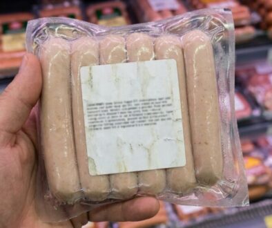 Hand holding a vacuum-sealed tray of pale pink sausages in a grocery store, with meat products visible on shelves in the background.