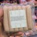 Hand holding a vacuum-sealed tray of pale pink sausages in a grocery store, with meat products visible on shelves in the background.