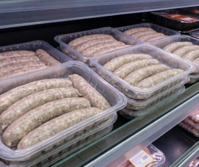 Plastic trays of raw sausages stacked on a store refrigerated shelf, ready for sale.
