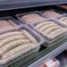 Plastic trays of raw sausages stacked on a store refrigerated shelf, ready for sale.