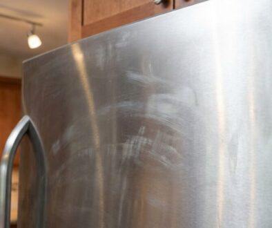 Stainless steel refrigerator door with smudges in a kitchen with wooden cabinets and track lighting in the background.
