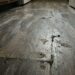 Low-angle view of a worn, dark wooden floor with scratches, stains, and peeling finish in a dim room.
