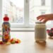 Child-friendly snack setup on a kitchen table: a colorful juice bottle, spilled gummy candies, a jar with a hand reaching to open it, plus berries and an apple nearby.