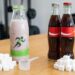 Berry smoothie in a bottle beside sugar cubes, a small scoop, and two cola bottles on a gym counter.