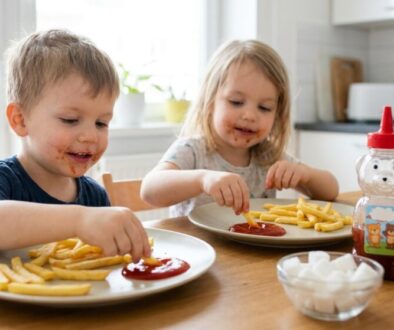 Two young children sit at a kitchen table, dipping fries in ketchup and smiling at their plates much eaten with messy faces.