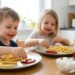 Two young children sit at a kitchen table, dipping fries in ketchup and smiling at their plates much eaten with messy faces.