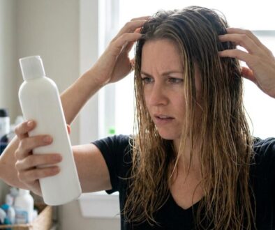 Woman with wet hair in a bathroom examines a large white bottle, touching her temples in confusion/concern.