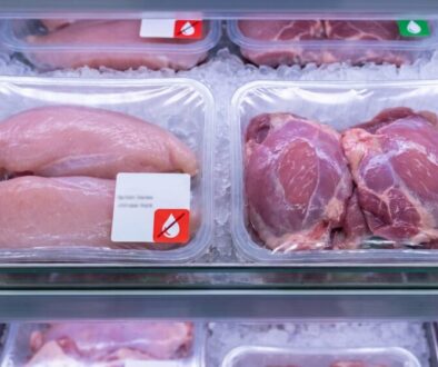 Tray of raw chicken breasts in a clear plastic package on a bed of ice in a supermarket display, with a small white label featuring a red droplet icon on the package.
