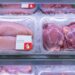 Tray of raw chicken breasts in a clear plastic package on a bed of ice in a supermarket display, with a small white label featuring a red droplet icon on the package.