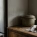 Rustic kitchen scene: a green ceramic pot on a wooden counter with a loaf of bread on a cloth by a window beside a stainless fridge.