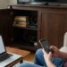 Woman on a couch holds a phone with a loading spinner while a laptop on the table shows a 'connection lost' message, in a living room with a cabinet and router cables.