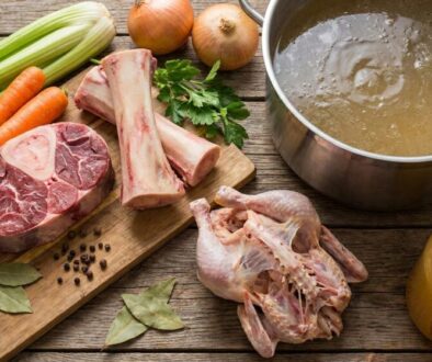 Assorted raw ingredients for soup on a wooden board: beef chop, lamb ribs, celery, carrots, onions, herbs, and a pot of stock nearby