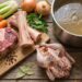 Assorted raw ingredients for soup on a wooden board: beef chop, lamb ribs, celery, carrots, onions, herbs, and a pot of stock nearby