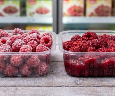 Two clear plastic containers on a wooden surface: frosted raspberries on the left and raspberry jam on the right, with a grocery freezer in the background.